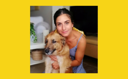 Woman in a blue patterned sleeveless dress sitting indoors and hugging a medium-sized tan and white dog, both looking toward the camera. The photo is encased by a yellow frame.